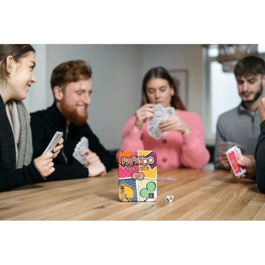 Boîte du jeu Papayoo placée au centre d’une table pendant une partie avec des joueurs en arrière-plan et un dé posé à côté.