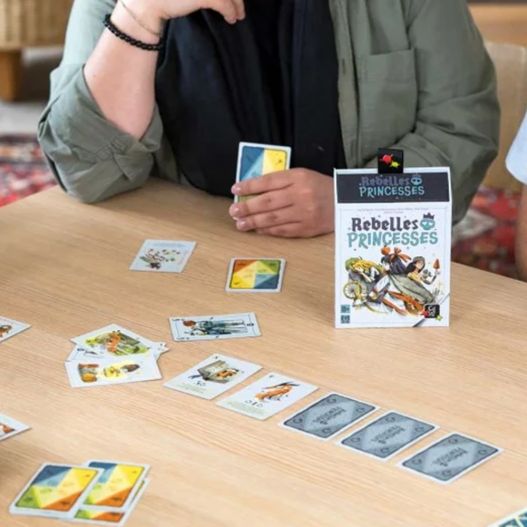 Boîte du jeu Rebelles Princesses sur une table, entourée de cartes en pleine partie.