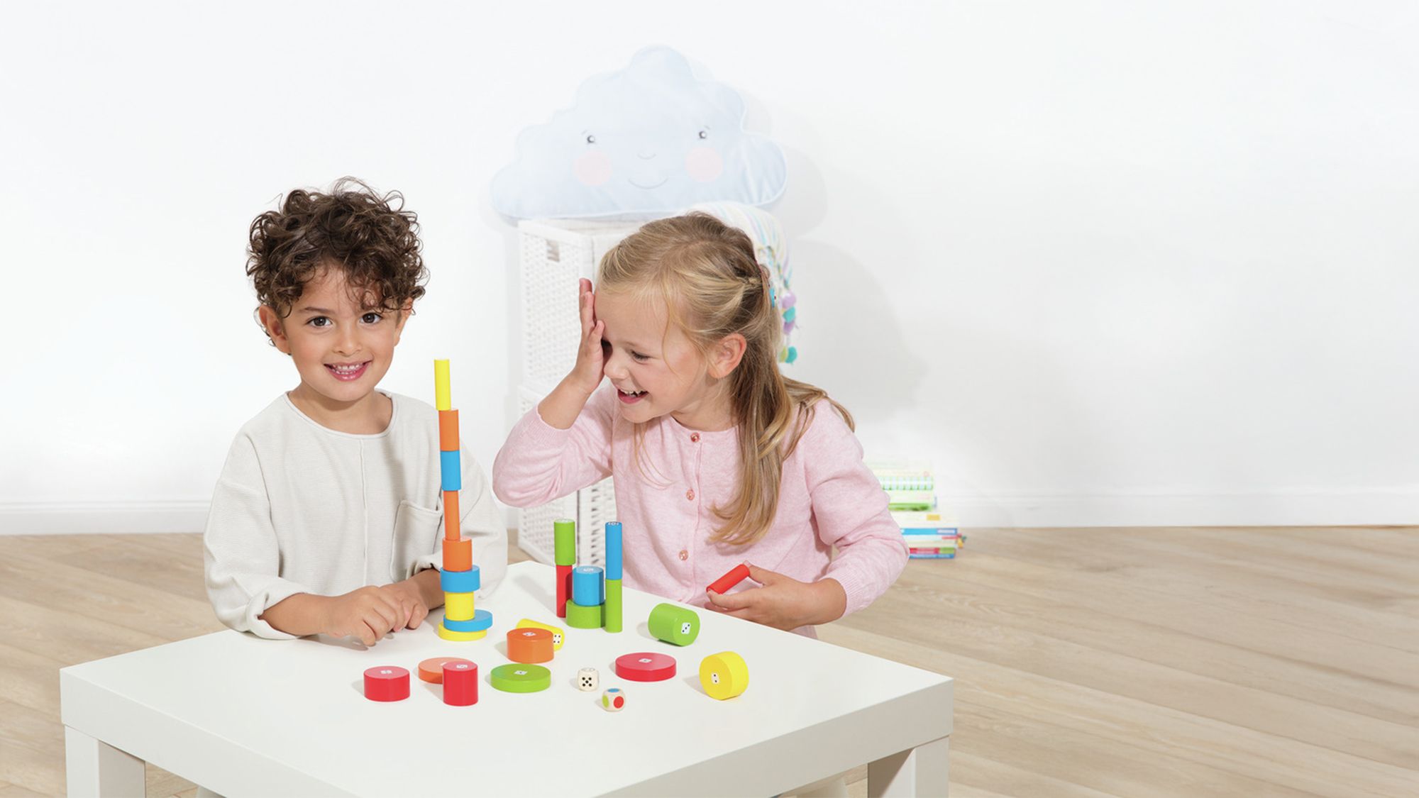 Deux enfants souriants jouant avec des blocs en bois colorés (bleu, vert, jaune, rouge) sur une table blanche dans une pièce lumineuse.