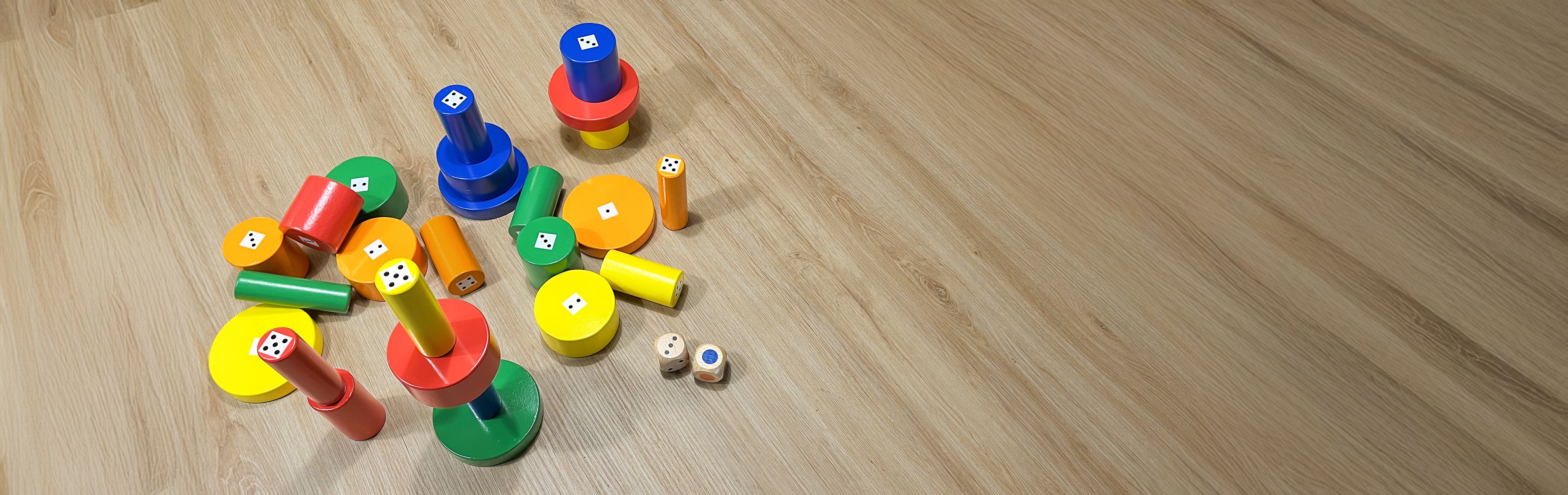 Jeux colorés en bois empilés, avec dés visibles et accessoires en forme de cylindres de couleur rouge, bleu, vert, jaune et orange sur un fond de sol en bois.