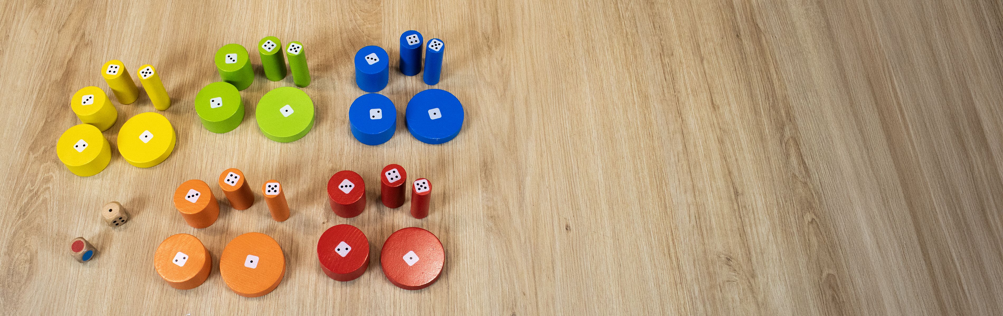 Jeux empilés en bois de différentes couleurs (jaune, bleu, vert, rouge, orange) avec des dés visibles, organisés en groupes sur un sol en bois.